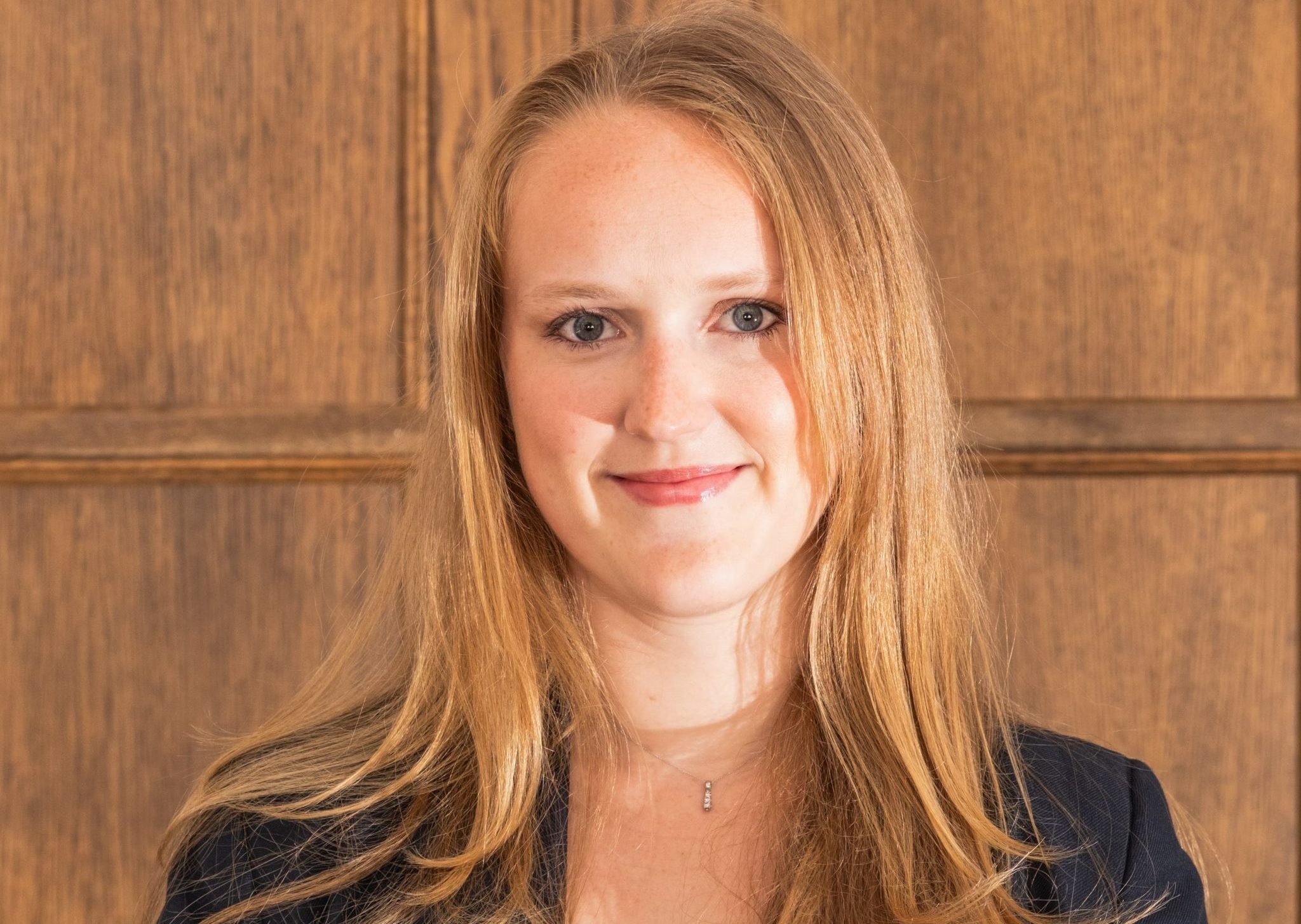A woman with shoulder length blonde hair wearing a dark blue blazer standing in front of a wooden panelling background.